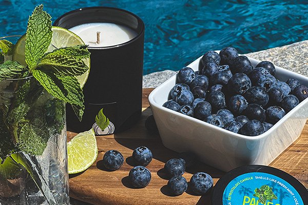 Sonicglow Pacific Beach candle styled with fresh mint, limes, blueberries and a wooden tray near water... representing multiple fragrance families.