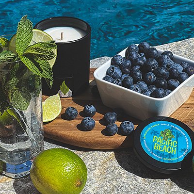 Sonicglow Pacific Beach candle styled with fresh mint, limes, blueberries and a wooden tray near water... representing multiple fragrance families.