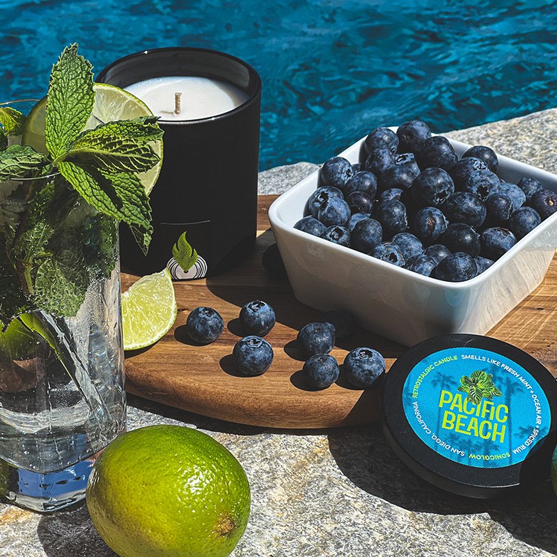 Sonicglow Pacific Beach candle styled with fresh mint, limes, blueberries and a wooden tray near water... representing multiple fragrance families.