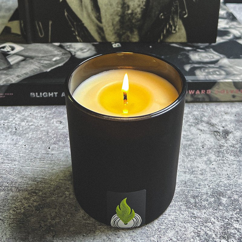 Lit Sonicglow candle with centered flame and partially melted wax pool, sitting on concrete with books in the background — showing proper burn for preventing candle tunneling.