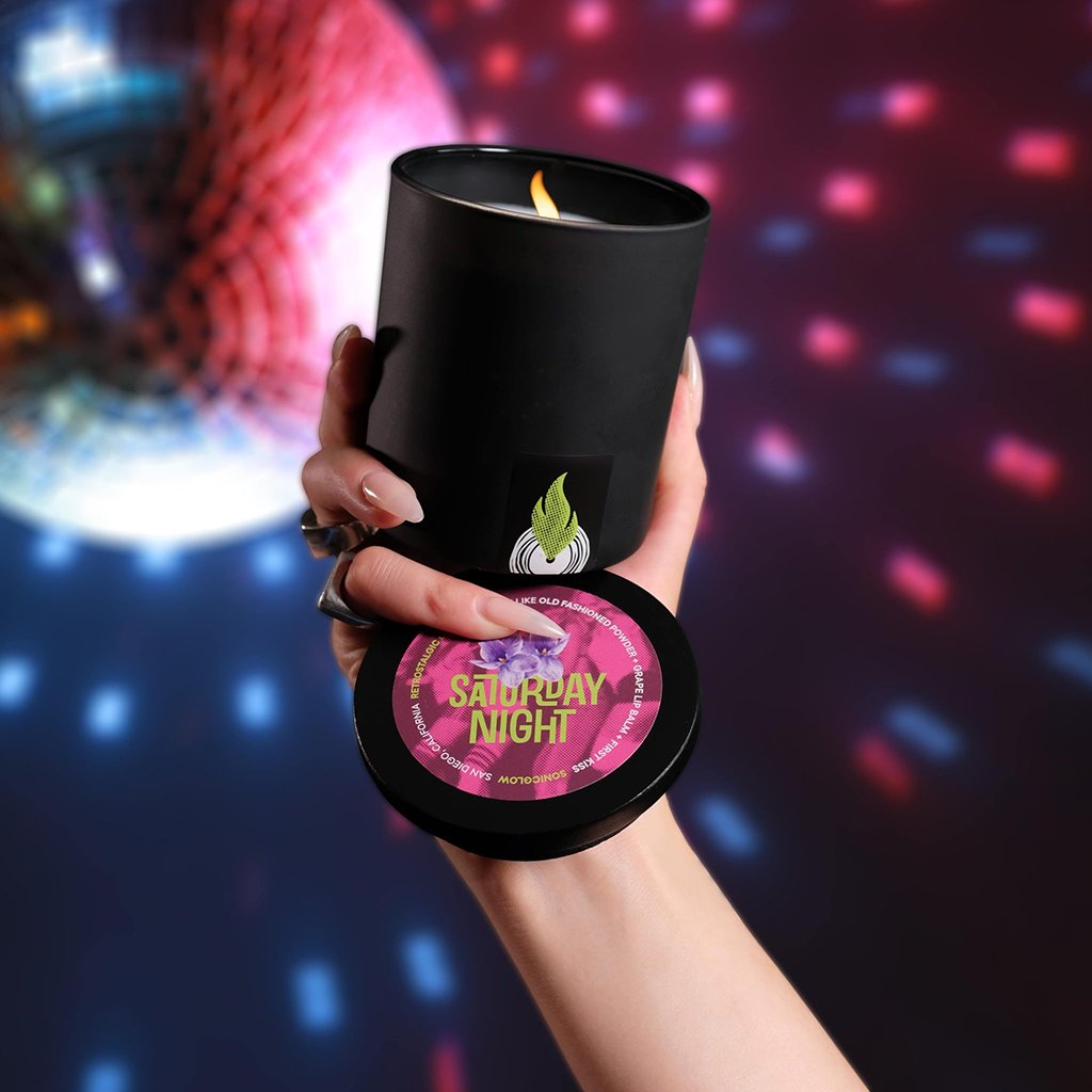 A hand holds a sleek black, ecoluxe Saturday Night scented candle from Sonicglow with a vibrant, retro label, against a blurred backdrop of a colorful disco ball dance floor lights