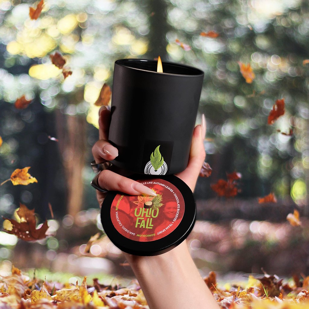 A hand holds a sleek black, ecoluxe Ohio Fall scented candle from Sonicglow with a warm, autumnal label design, lit and casting a cozy glow against a bokeh background of a forest with falling leaves