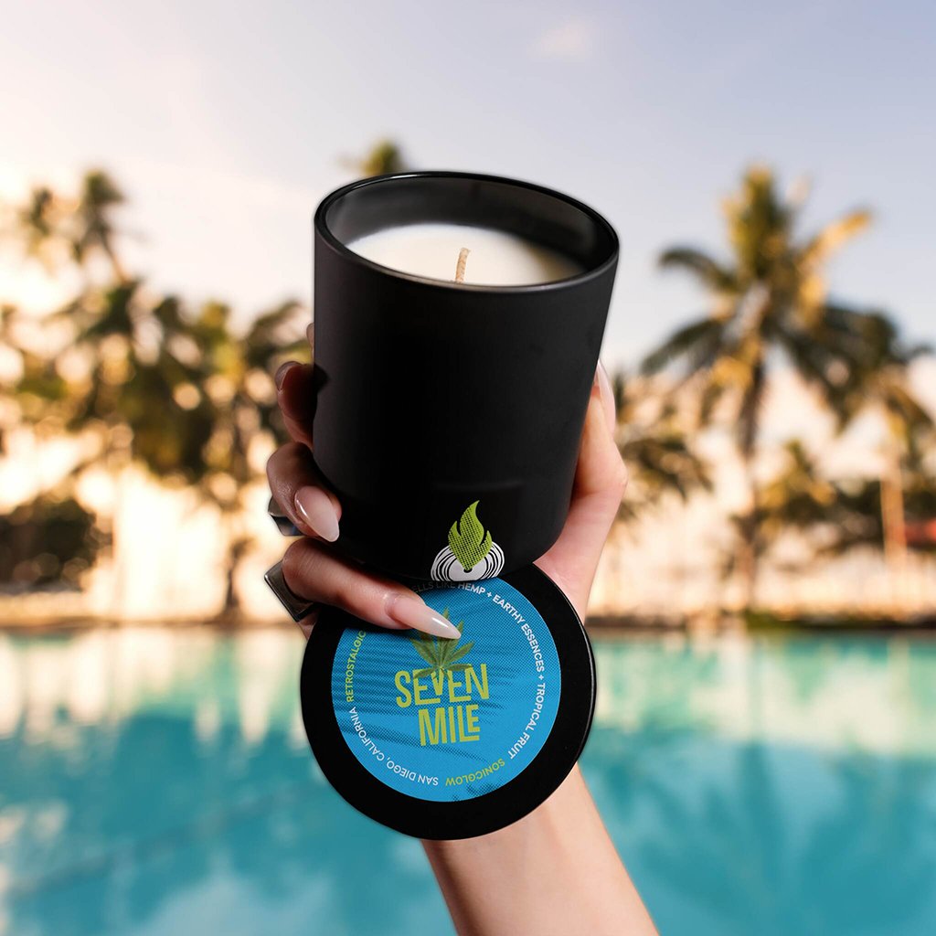 A hand holds a sleek black, ecoluxe Seven Mile candle from Sonicglow with a bright blue label featuring tropical design elements, poised against a serene, blurred poolside backdrop with palm trees